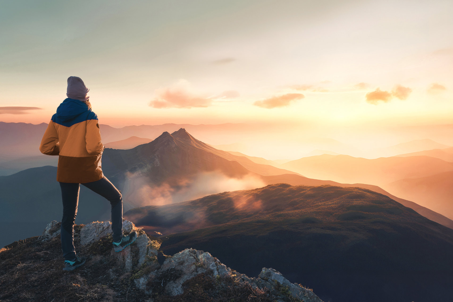 Eine weibliche Person von hinten steht auf einem felsigen Berggipfel und blickt auf eine weite, neblige Bergkette bei Sonnenuntergang. Sie trägt eine senfgelbe, braune und blaue Jacke, dunkle Hosen und eine Mütze. Das Licht des Sonnenuntergangs ist warm und orange.